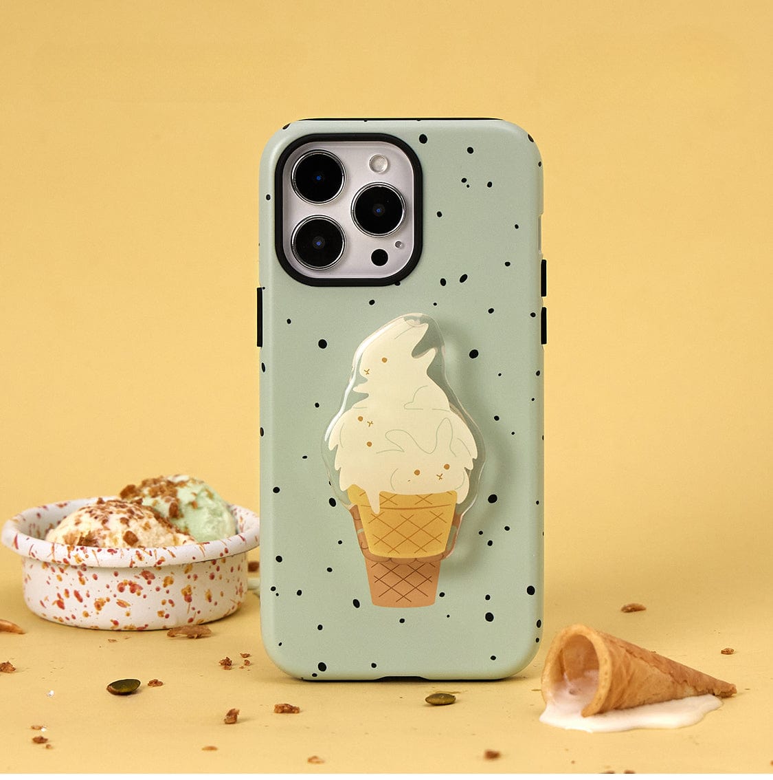 The Mint Ice Cream MagSafe iPhone 16 Pro Max Case, featuring a mint-green design with black speckles and a cartoon ice cream cone, is displayed against a yellow background. Nearby, a toppled ice cream cone and a bowl of mint chocolate chip ice cream with toppings enhance the sweet treat theme.