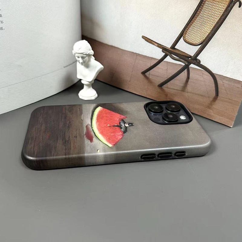 A Quirky Watermelon & Miniature Hero iPhone 16 Pro Max Case - Fun and Unique Design, Ultra-Slim, Full Protection is placed on a gray surface. Nearby are a miniature bust sculpture, an open book, and a wicker chair against a wall.