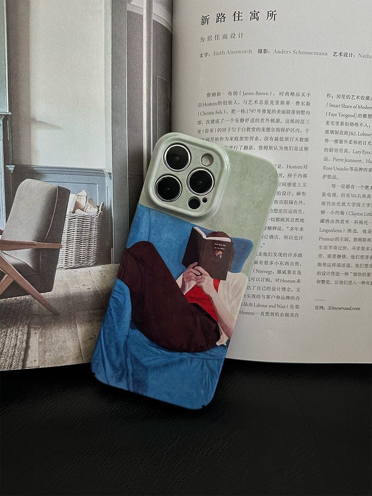 A smartphone encased in the Relaxed Reader iPhone 16 Pro Max Case - Artistic and Protective, adorned with an illustration of a person reading a book while sitting on a blue chair, is placed on an open magazine. The phone stands upright, partially obscuring the text and images on the magazine page.