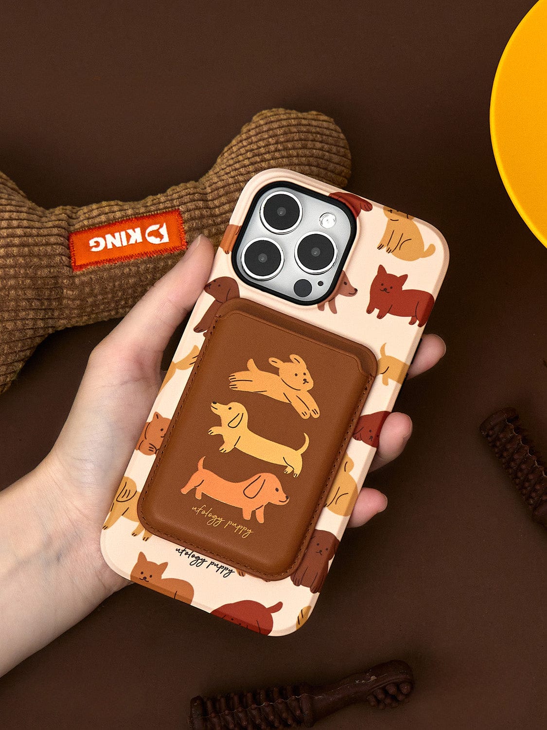 Hand holding a Puppy Playtime MagSafe iPhone 16 Pro Max Case with cute dog illustrations and an integrated stand, featuring a brown cardholder. A brown textured dog toy and a yellow object are visible in the background.