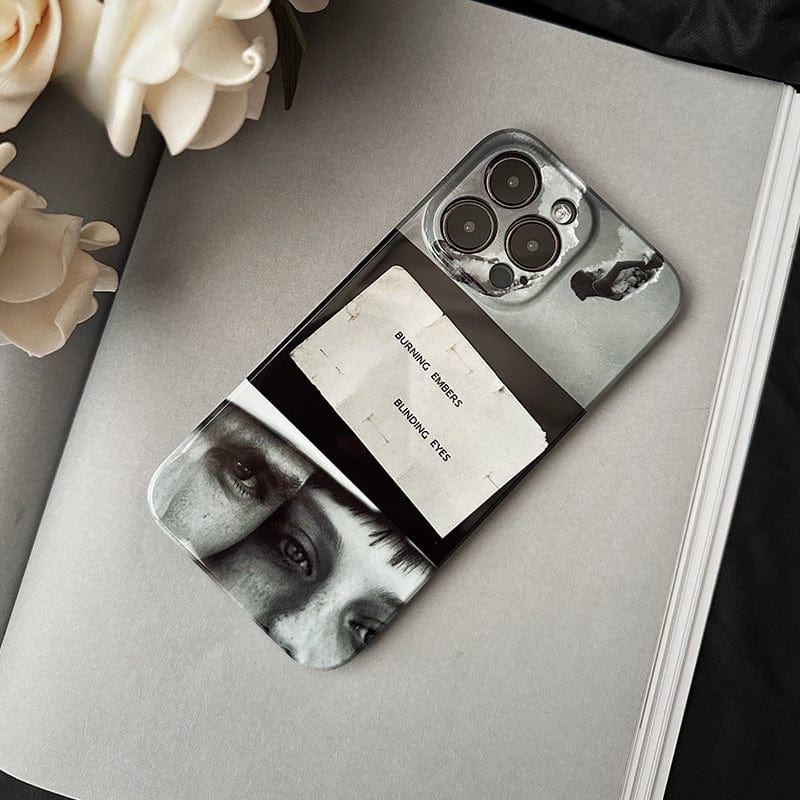 An iPhone 16 Pro Max, adorned in a Moody Collage case featuring an artistic black and white design with close-ups of eyes and abstract imagery, lies on an open book beside white flowers. The case also includes the thought-provoking quote, "BURNING DESIRE, BURNING EYES," while the phone itself boasts three camera lenses.