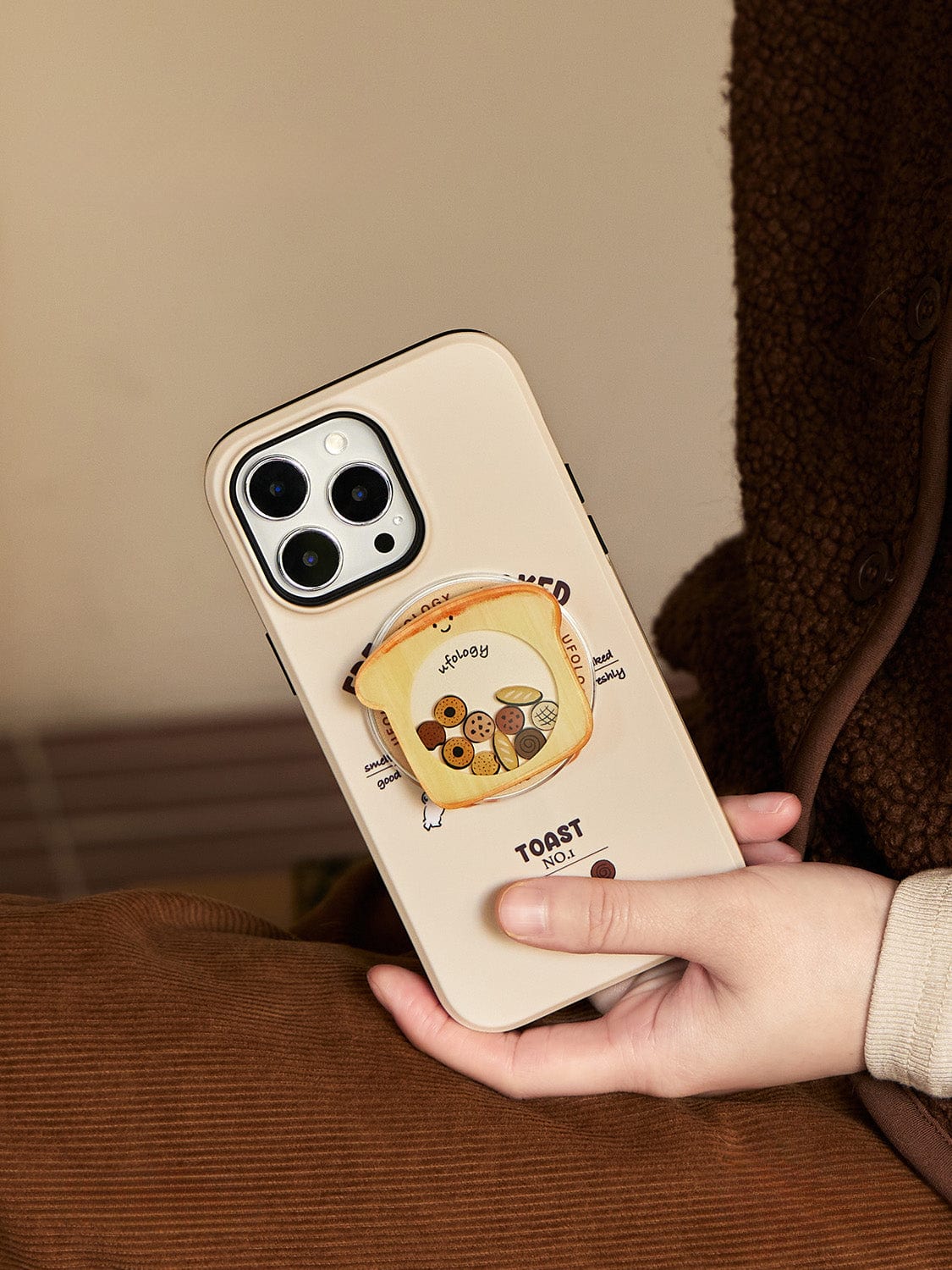 A person holds a Morning Toast MagSafe iPhone 16 Pro Max Case, which features an illustration of a smiling toast character with "Happy" written on it, surrounded by various types of bread. The person's outfit includes a brown, textured fabric, possibly a jacket or sweater.