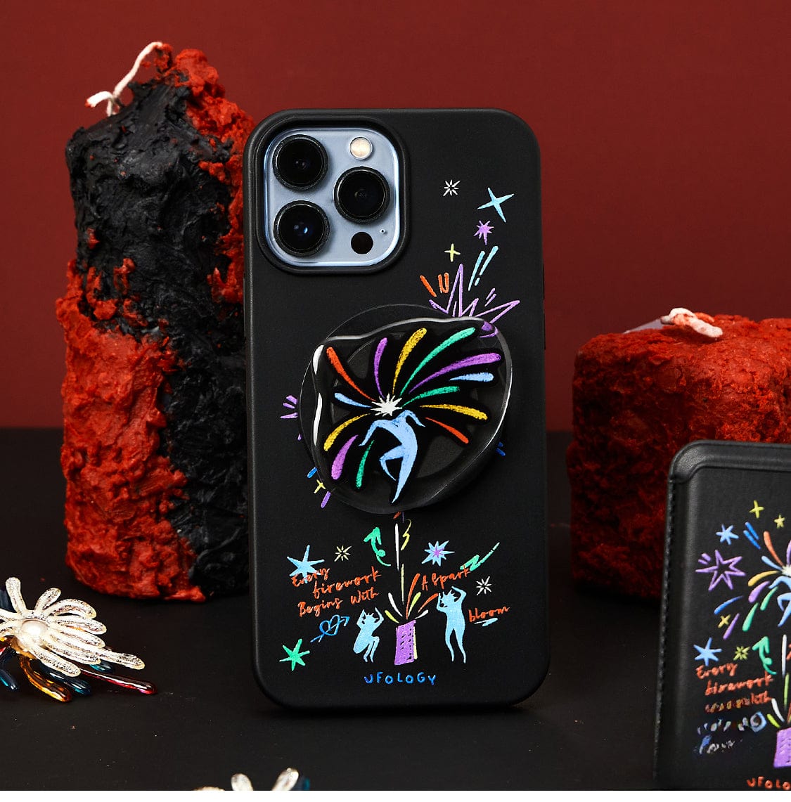 A close-up of the Firework Fantasy MagSafe iPhone 16 Pro Max Case, showcasing its vibrant celebration design featuring whimsical dancing figures, stars, and animals set against a backdrop of red and black sculptures. The case includes a pop socket adorned with a dynamic burst pattern.