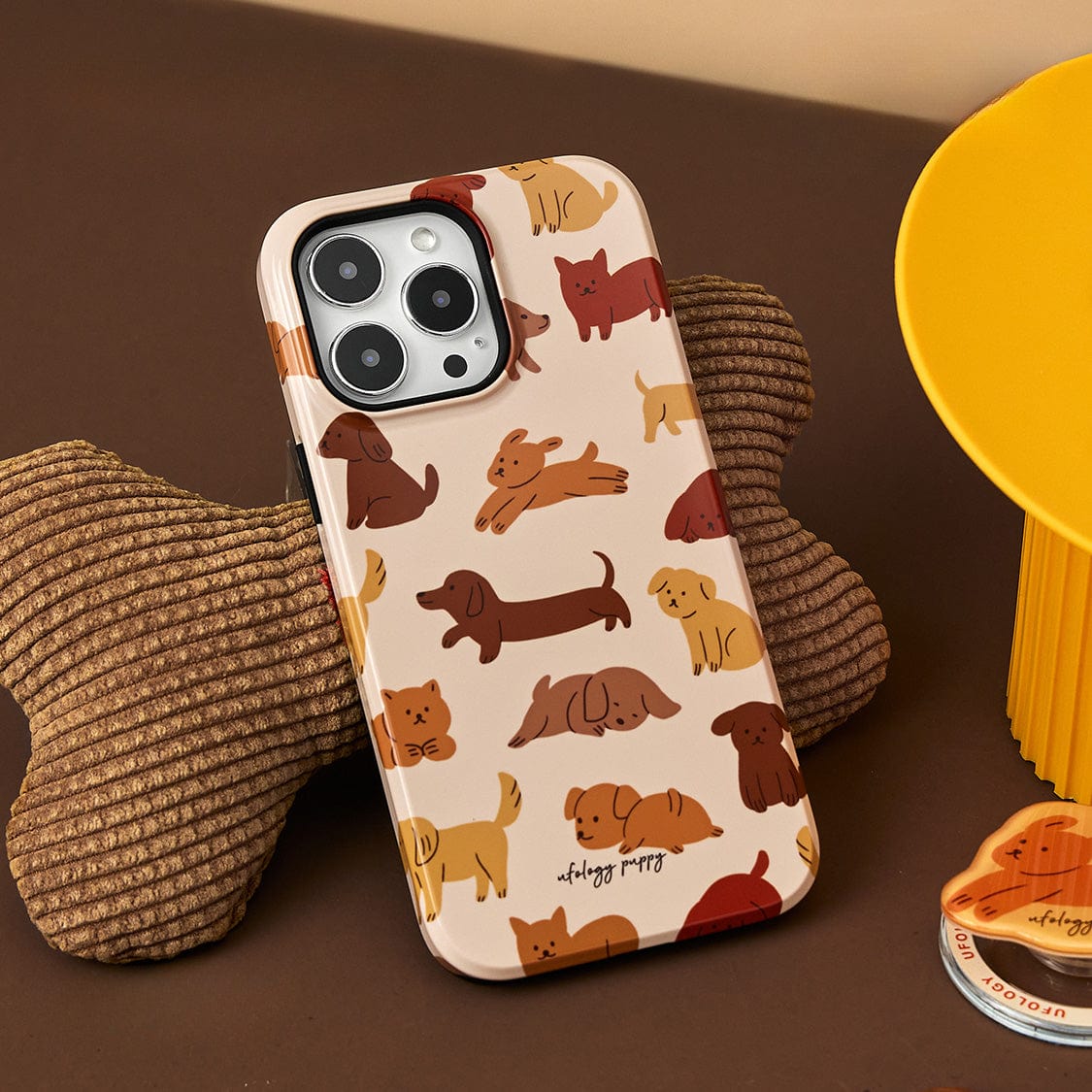 Puppy Playtime MagSafe iPhone 16 Pro Max Case, showcasing cute cartoon dog illustrations, resting on a brown, bone-shaped cushion beside a yellow object.