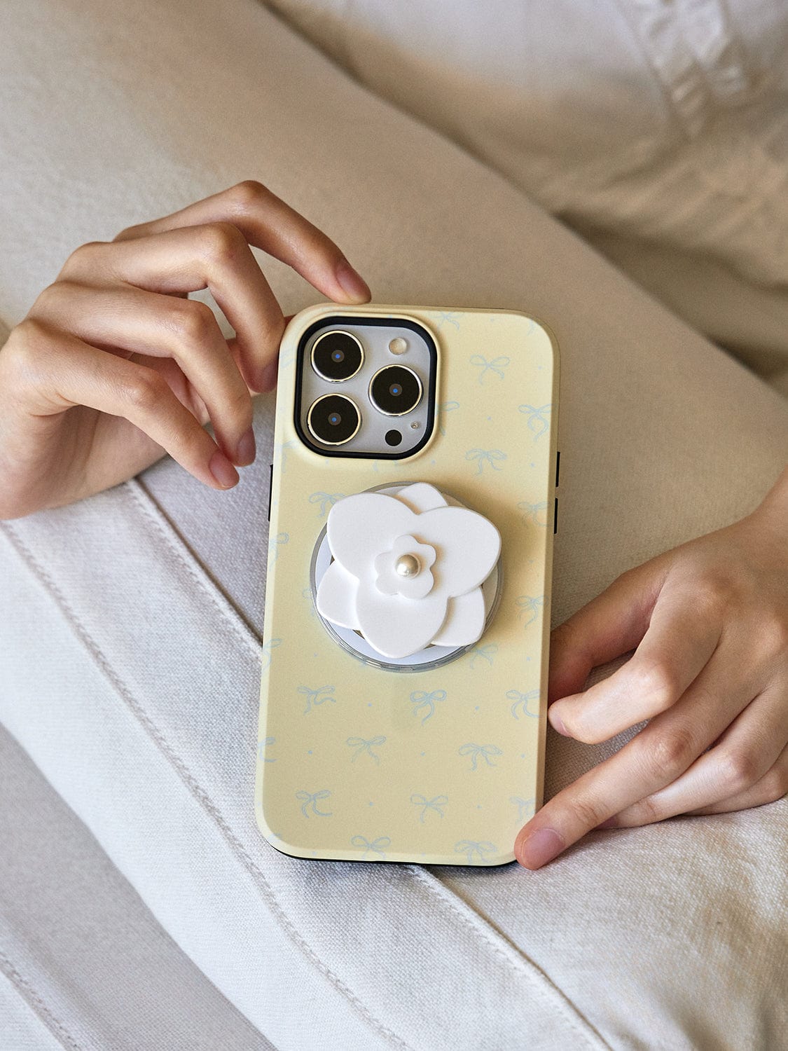 Close-up of a person holding an Elegant Blossom MagSafe iPhone 16 Pro Max Case, showcasing its chic floral design with a pearl flower stand. The person is seated on a light-colored couch.