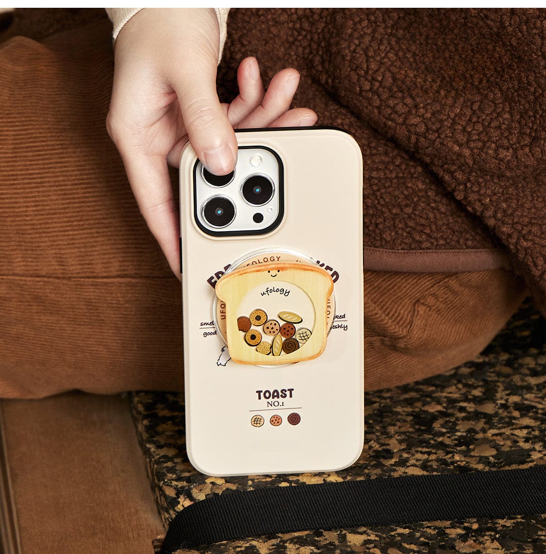 A person is holding a Morning Toast MagSafe iPhone 16 Pro Max Case, which features a cream-colored cover adorned with a cute breakfast design, including a toast stand labeled "toast" that can store small items like earbuds. The background showcases a brown textured surface and the leg of someone dressed in brown.