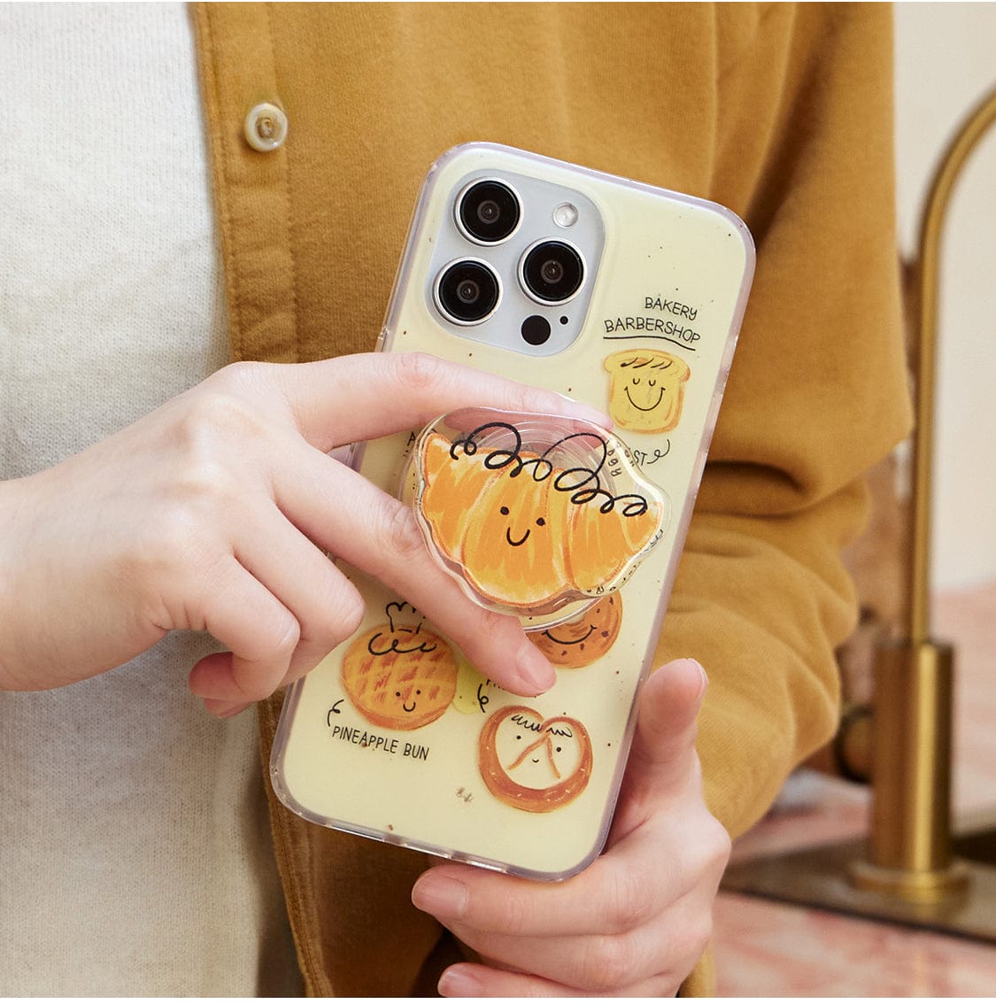 A person is holding the Bakery Friends MagSafe iPhone 16 Pro Max Case, which features a cute decorative pattern of illustrated baked goods, including a smiling pineapple bun. The light-colored phone case includes an all-inclusive protective cover and a bun stand with the same pineapple bun design. The person is wearing a mustard-colored top.