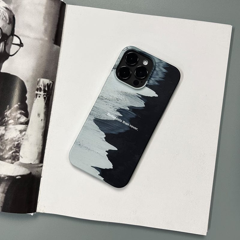 A smartphone adorned with the Monochrome Waves iPhone 16 Pro Max Case, which boasts a minimalist black and white abstract design and the quote "Better Late Than Never," rests on an open book displaying grayscale images. The book is placed on a gray surface.