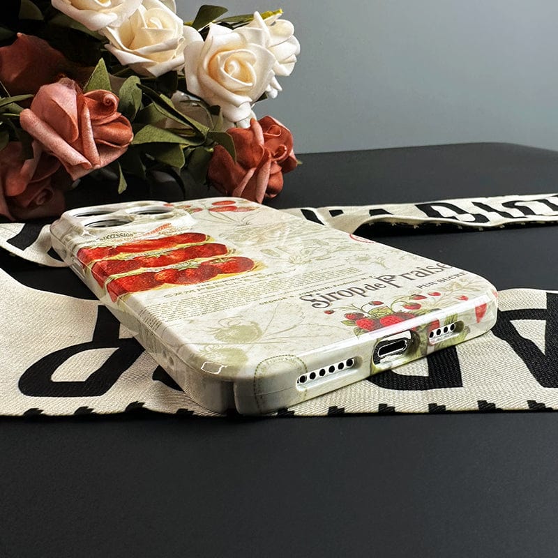 A smartphone with a Vintage Strawberry Print iPhone 16 Pro Max Case - Elegant and Protective, showcasing red and green patterns with vintage graphics, sits on a black surface. Behind it, there's a bouquet of white and pink roses arranged decoratively on beige and black fabric.