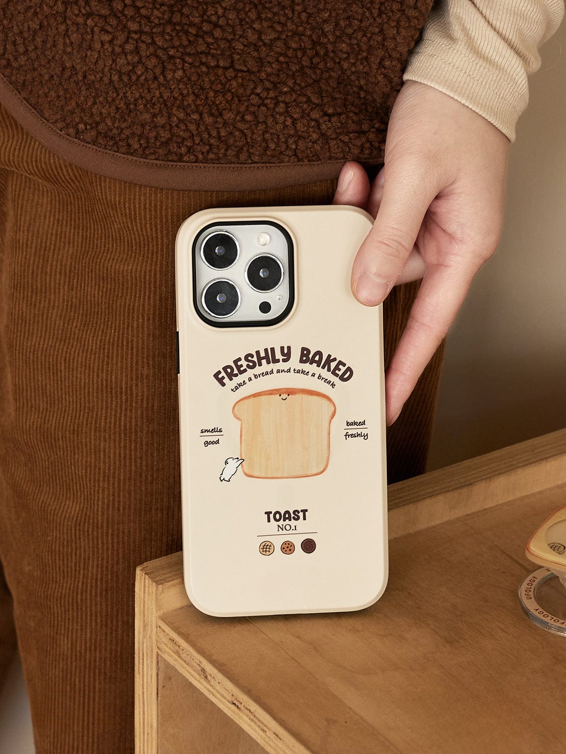 A person holds the Morning Toast MagSafe iPhone 16 Pro Max Case, featuring a cute breakfast design with text that reads "Freshly Baked: Grab a Bread and Take a Break. Toast No. 1." The beige case showcases a smiling toast with arms. The person is wearing brown corduroy pants and a cozy outfit.