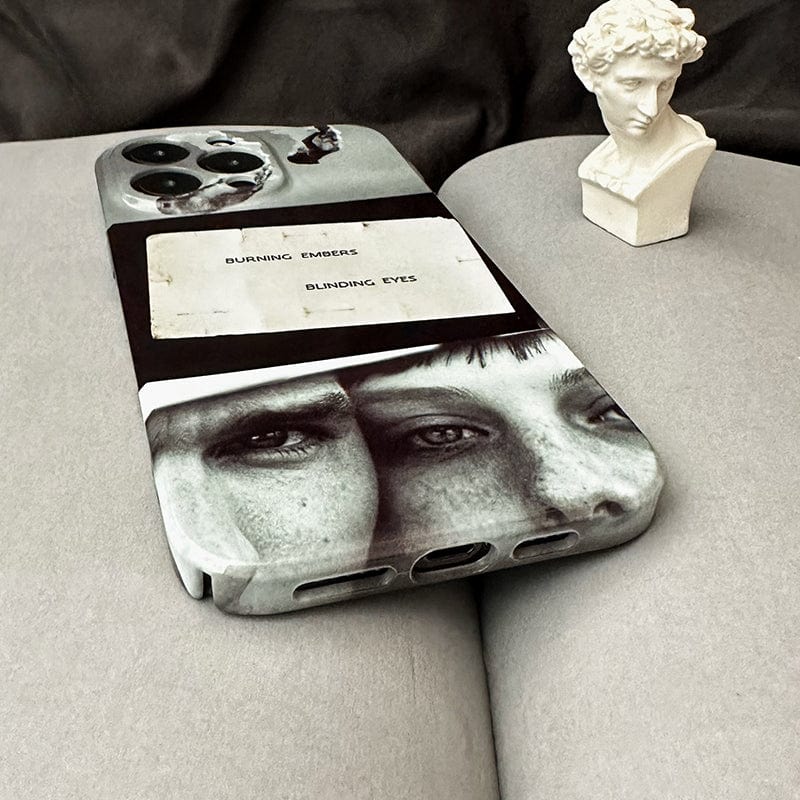 An iPhone 16 Pro Max lies on a beige surface, protected by the Moody Collage iPhone 16 Pro Max Case, which features an artistic black and white design with images of two portraits. The case is adorned with the thought-provoking quote "BURNING EMBERS BLINDING EYES." Next to the phone is a small, white bust sculpture of a classical figure.