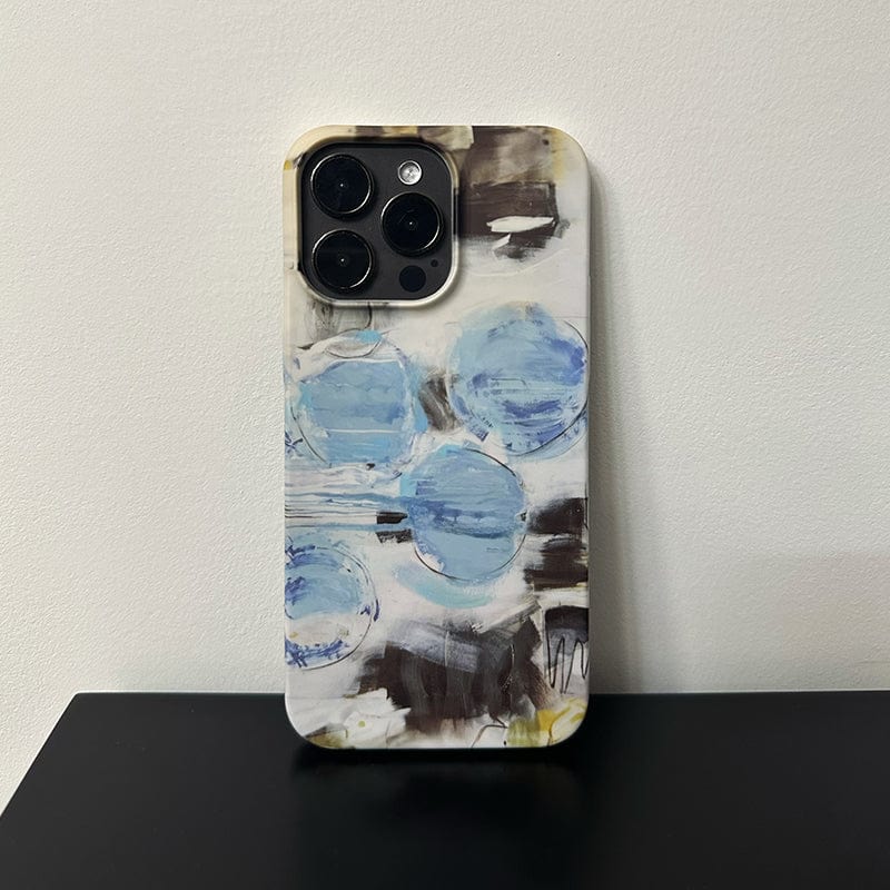 An iPhone 16 Pro Max with the Abstract Blue Circles Case, featuring a modern art-inspired design in shades of blue, black, and white—complete with overlapping circles and various brush strokes—is resting upright on a black surface against a white wall.