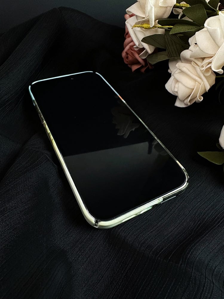 A smartphone with a reflective screen and a Whimsical Cat Dream iPhone 16 Pro Max case lies on a textured black fabric. In the background, there is a bouquet of artificial flowers with white and pink blossoms, and green leaves.