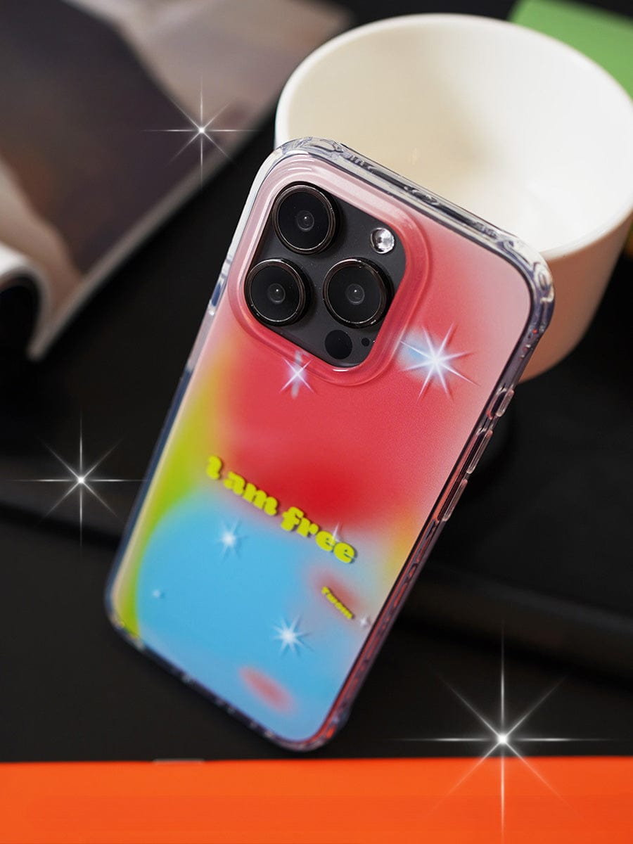 A smartphone with an "I Am Free Gradient iPhone 16 Pro Max Case" sits on a table. The case features a vibrant gradient of pink, yellow, and blue along with the text "I am free" and several star-like sparkles. In the background, there is a white cup and a blurred piece of fabric.