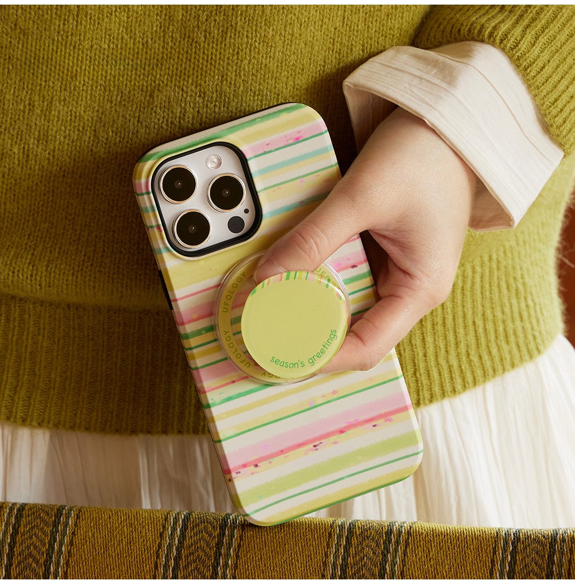 A person holds an iPhone with a Festive Stripes MagSafe iPhone 16 Pro Max Case, featuring a cheerful holiday design in green and pink stripes. The case includes a circular grip accessory with the words "season's greetings" written on it. They are dressed in a green sweater over a white shirt.