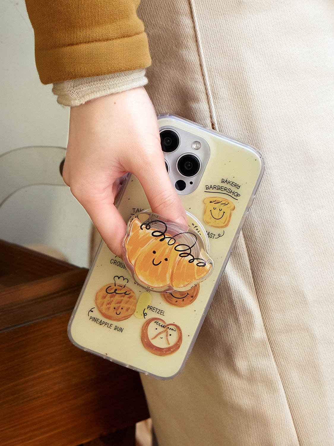 A person wearing a brown and beige outfit is holding a smartphone with the "Bakery Friends MagSafe iPhone 16 Pro Max Case." The clear case, featuring smiley-faced buns and pastries like croissants, pretzels, and pineapple buns, also has a circular smiling bun-shaped pop socket attached to the back for added convenience.