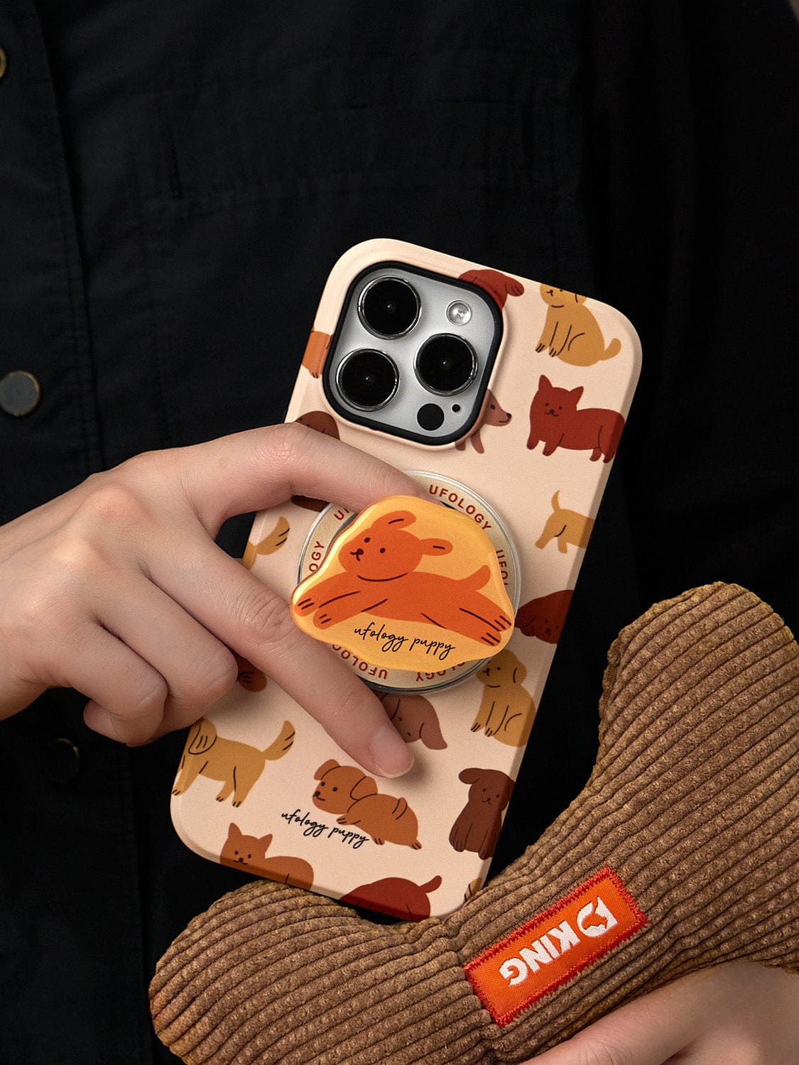 A person is holding a smartphone encased in a Puppy Playtime MagSafe iPhone 16 Pro Max Case, featuring an adorable dog design and an integrated stand. The hand also grips a textured toy labeled "KING" in the background.