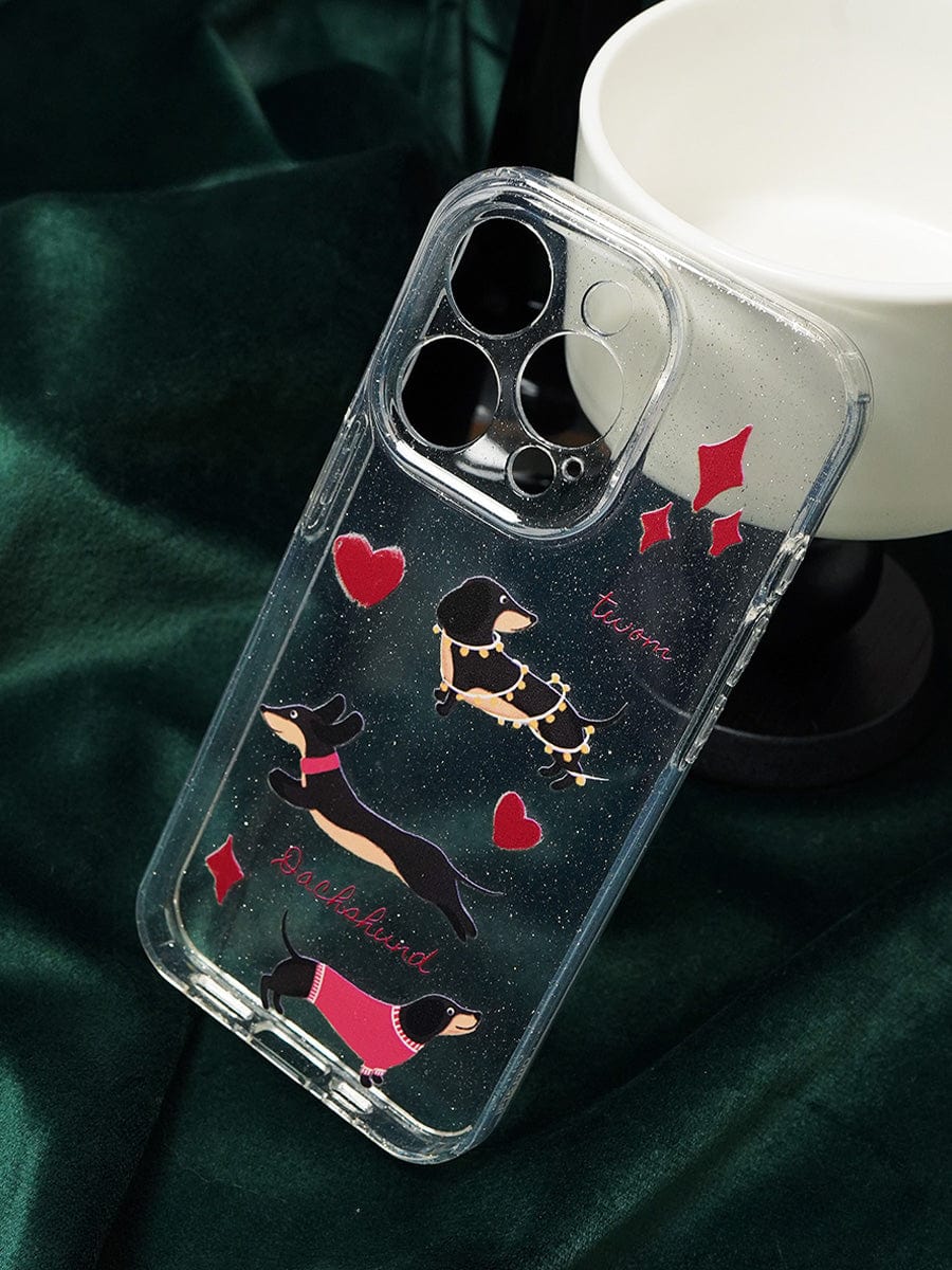 A clear sparkle-design "Dachshund Love iPhone 16 Pro Max Case" featuring illustrations of three dachshund dogs in colorful outfits, accented with red hearts and the words "Dachshund" and "Love." The cute all-inclusive protective cover is showcased on a dark green fabric background, with a partially visible white object nearby.