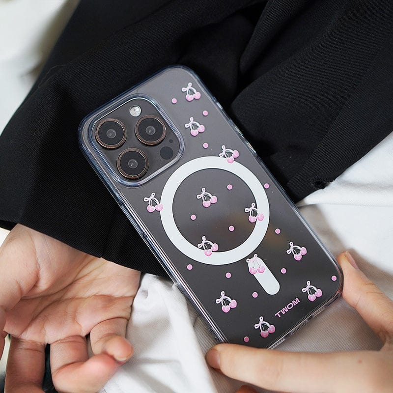 A close-up view of a smartphone in a person's hand, showcasing the Cherry Blossom MagSafe iPhone 16 Pro Max Case with an adorable 3D cherry design and a circular white area on the back, suggesting support for MagSafe charging. The person's hand is resting on black fabric.