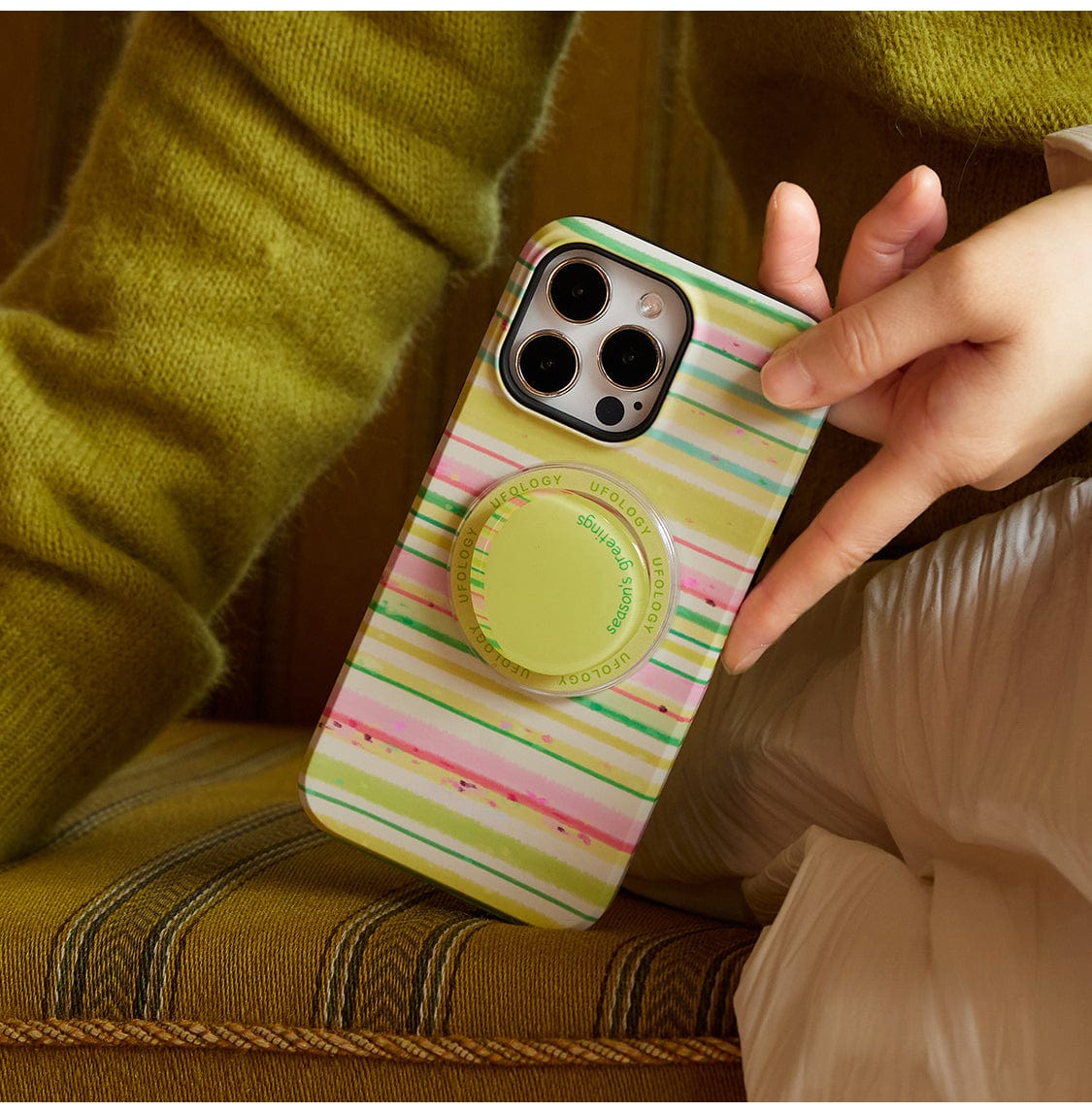 An individual, dressed in a green sweater and white bottoms, sits on a striped green and brown cushion while holding the Festive Stripes MagSafe iPhone 16 Pro Max Case. This cheerful holiday design boasts charming striped patterns in green, pink, and yellow along with a convenient circular grip featuring inscriptions.