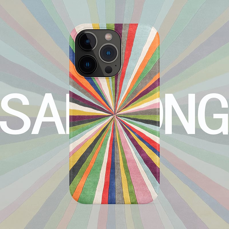 The Vibrant Burst iPhone 16 Pro Max Case features a colorful radiant stripes design, where a multicolored radial pattern of red, green, yellow, orange, pink, and blue stripes converge at a center point. The striking design fans out against the background for full protection. Note that the word “SAMSUNG” is partially visible behind the case.