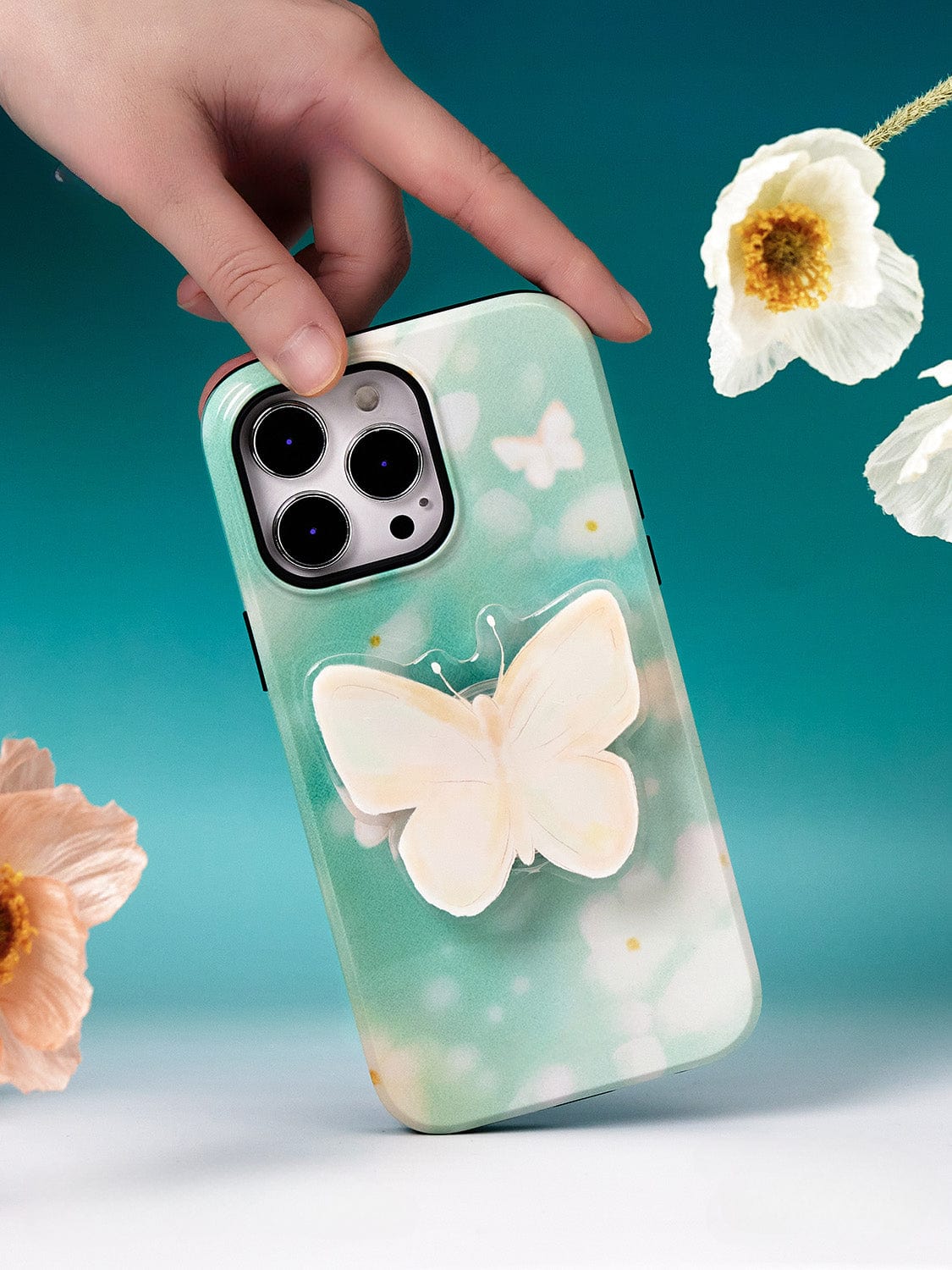 A hand holds a smartphone with a Butterfly Blossom MagSafe iPhone 16 Pro Max Case against a teal background adorned with white and orange flowers.