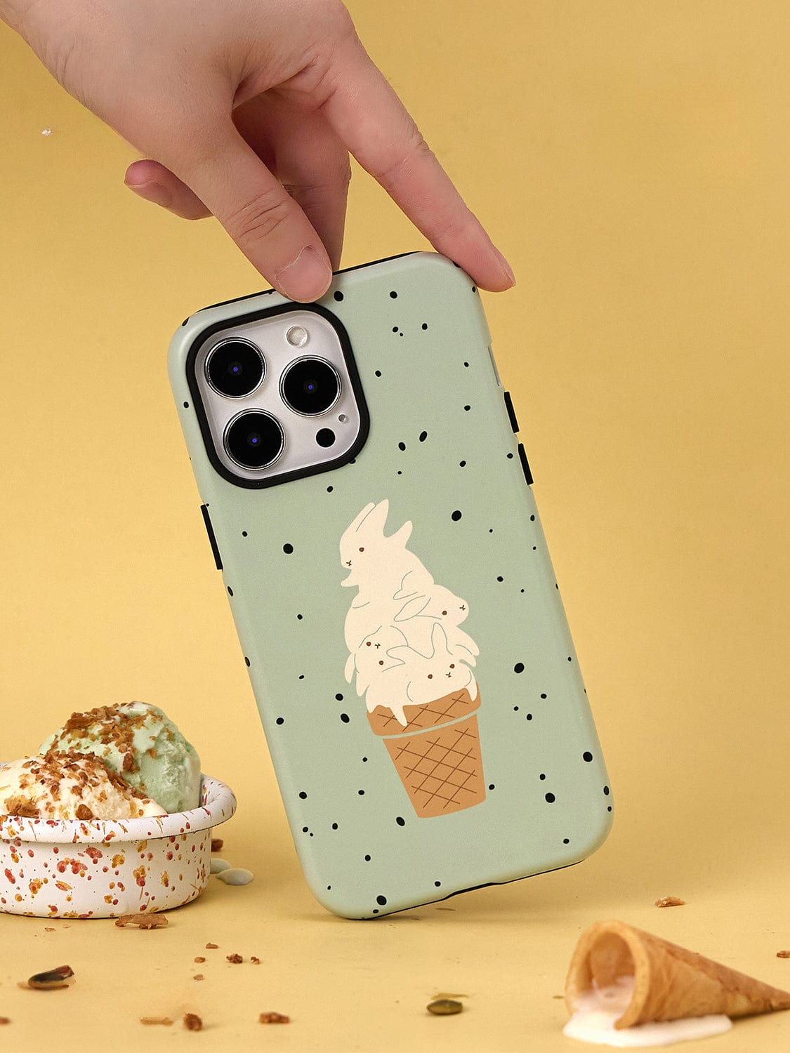 A hand holds the Mint Ice Cream MagSafe iPhone 16 Pro Max Case | Sweet Treat Design with Stand, featuring an illustration of bunnies perched on an ice cream cone. The background is yellow, and beneath the phone case lies a bowl with partially melted ice cream and a spilled cone with some scattered crumbs.