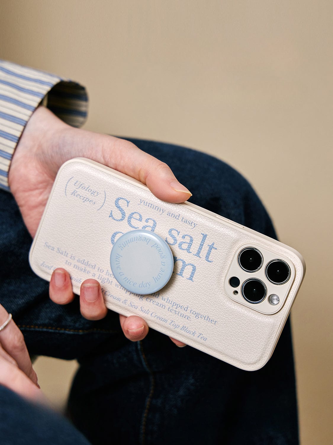 A person is holding an iPhone 16 Pro Max with a "Sea Salt Cream" MagSafe case, featuring a minimalist textured design, and a blue phone grip.