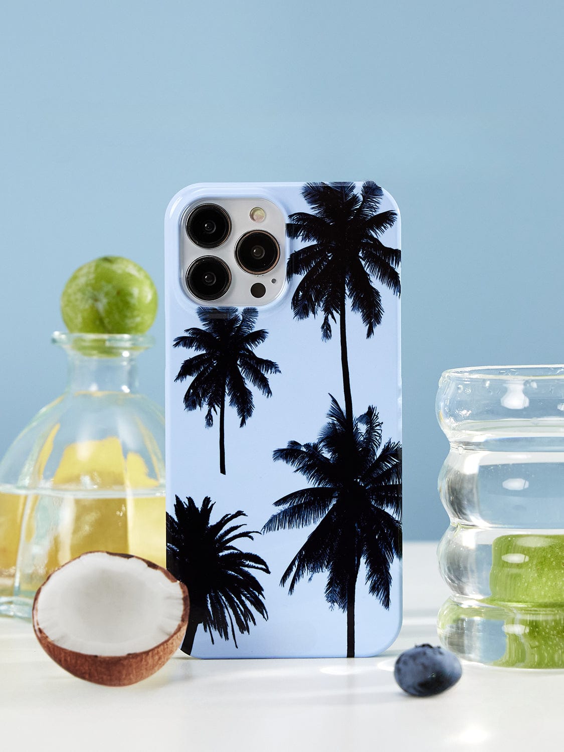 A sleek Tropical Escape MagSafe iPhone 16 Pro Max Case, showcasing black palm trees against a light blue background, is placed on a white surface. Surrounding the phone are a lime, a coconut, a glass pitcher with yellow liquid, a glass of water, and a single blueberry.