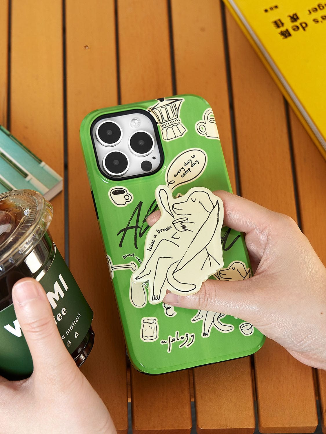 A hand is holding the Lazy Day Elephant MagSafe iPhone 16 Pro Max Case, featuring a playful green design with various illustrations, including an elephant and words, next to another hand holding a plastic cup. A yellow book and other items are seen in the background.