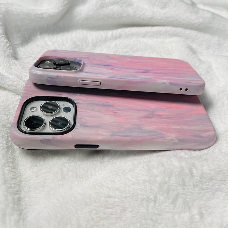 Two smartphones with pastel pink and purple painted cases are placed on a soft, white, textured fabric. The larger Pastel Brushstrokes iPhone 16 Pro Max Case - Soft Artistic Design with Full Protection is on the bottom, and the smaller one is on top, both showing their rear camera lenses.