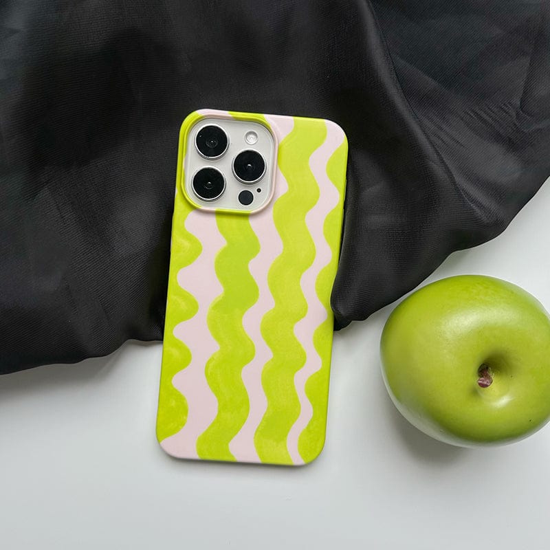 A smartphone encased in a Vibrant Wavy Line iPhone 16 Pro Max Case - Bold Lime Green & Cream Design, Ultra-Slim, Protective Cover is placed on a dark fabric background. Next to the phone, a green apple rests on a pristine white surface.