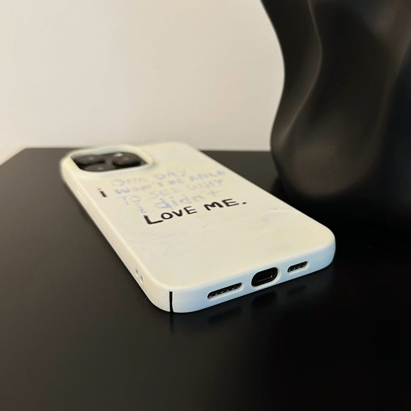 A white iPhone 16 Pro Max with a Minimalist Inspirational Quote case, featuring partially visible text reading "LOVE ME," lies flat on a black surface. Beside it is a dark object with a smooth, curving shape, captured in a close-up image focusing on the bottom and backside of the phone.