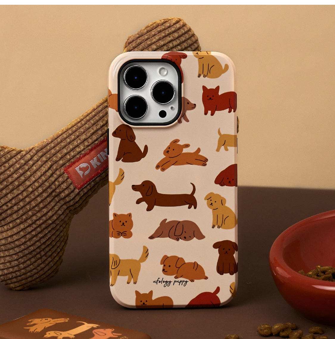 The Puppy Playtime MagSafe iPhone 16 Pro Max Case features a cute dog design with illustrations of various dog breeds in shades of brown, beige, and red. A textured dog bone toy and a red bowl filled with kibble are in the background, adding a playful touch to this all-inclusive protective cover with stand.