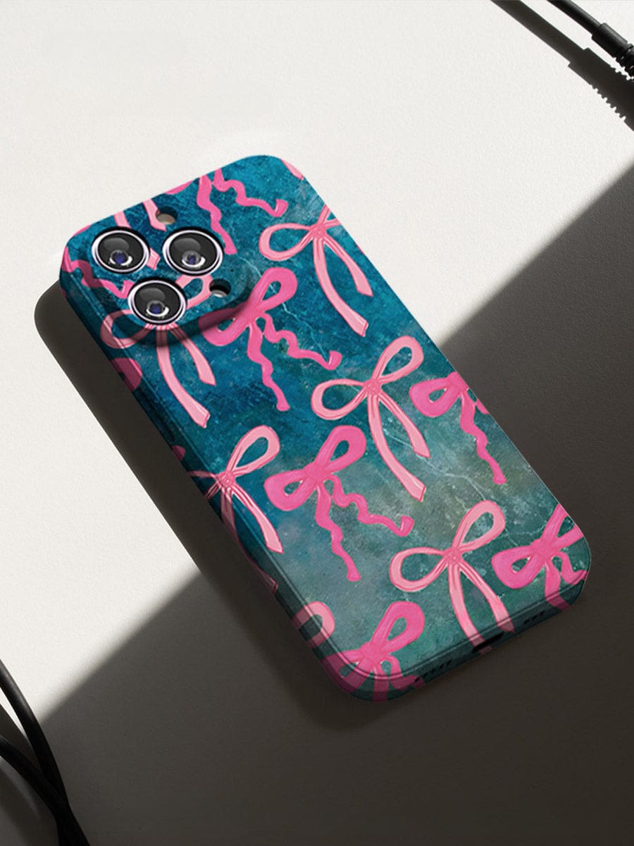 A smartphone adorned with the Pink Ribbon Charm iPhone 16 Pro Max Case, featuring an artistic bow design with pink bows on a blue and green background, is placed on a white surface. Light and shadow create a diagonal line across the image, while black cables are partially visible at the edges.