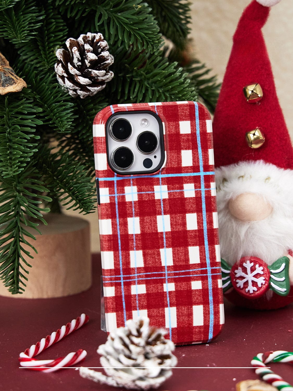 A Festive Knitted MagSafe iPhone 16 Pro Max Case, adorned with a holiday plaid design and featuring Santa and tree stands, sits against a festive holiday backdrop complete with a decorated evergreen branch, a small Santa figure in a red hat, pinecones, and candy canes.