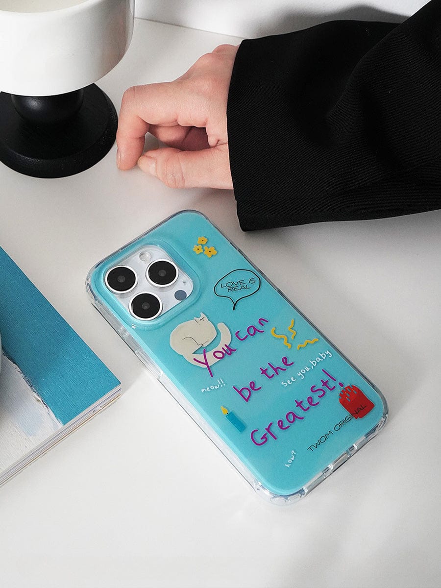 A hand reaches for the "You Can Be the Greatest iPhone 16 Pro Max Case" with a fun motivational design and colorful drawings, resting on a white table beside a book and a lamp.