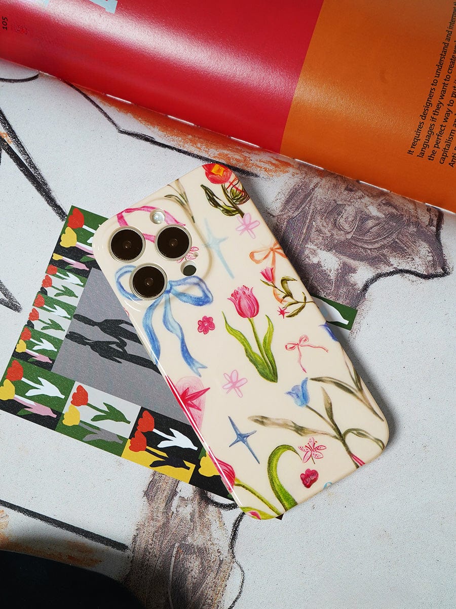 A Floral Dreams iPhone 16 Pro Max Case with a colorful botanical design cradles the smartphone, resting on an illustrated surface with a partly visible magazine in the background.