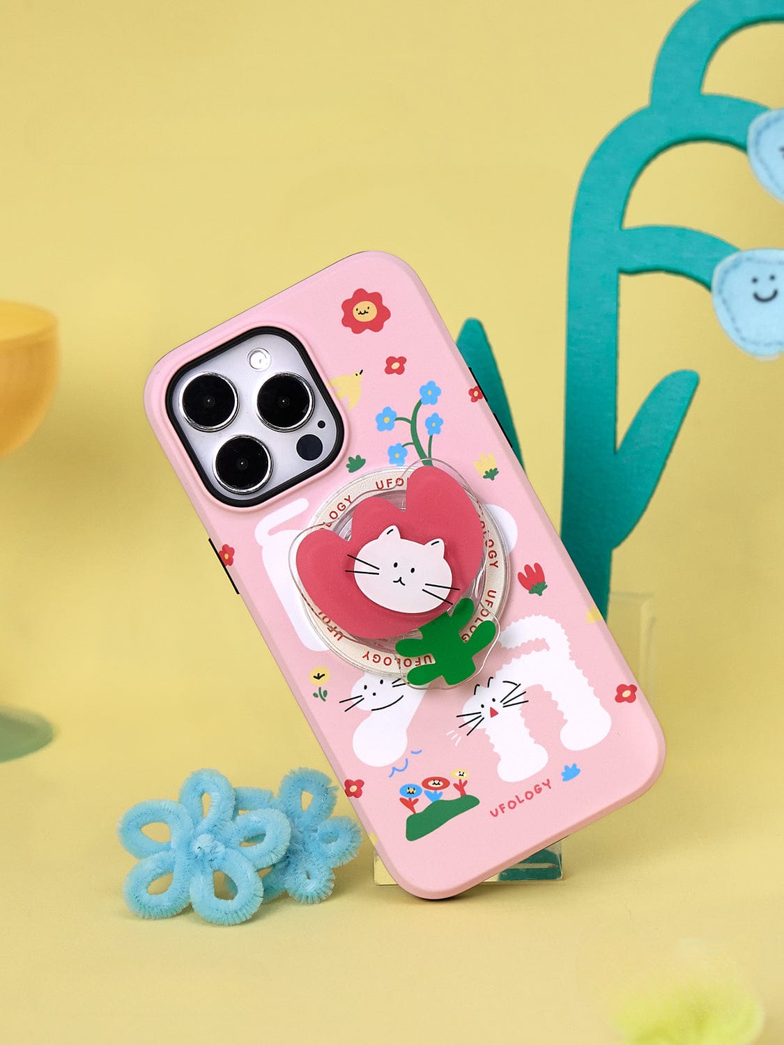 A Spring Kitty MagSafe iPhone 16 Pro Max Case featuring a playful floral design and cat stand is displayed upright against a yellow background with whimsical decorations.