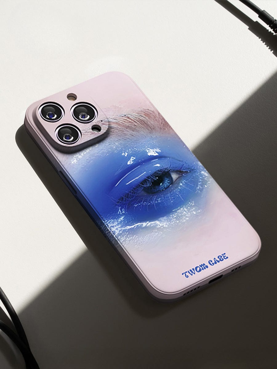 A white case for the iPhone 16 Pro Max, featuring an artistic design of a closed eye adorned with blue eyeshadow, and small black text near the bottom reading "TWOM CASE." The phone rests on a white surface with part of a cable visible nearby. This Dreamy Blue Eye case offers surreal art design and elegant, all-inclusive protection.