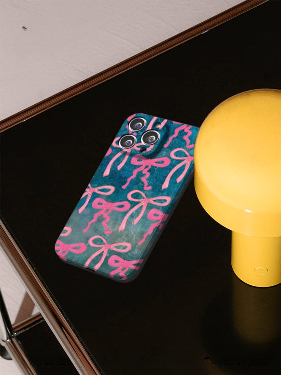 A smartphone with a Pink Ribbon Charm iPhone 16 Pro Max Case featuring an artistic bow design sits on a dark, glossy surface next to a yellow cylindrical object. The background is neutral, with a wall visible in the upper part of the image.