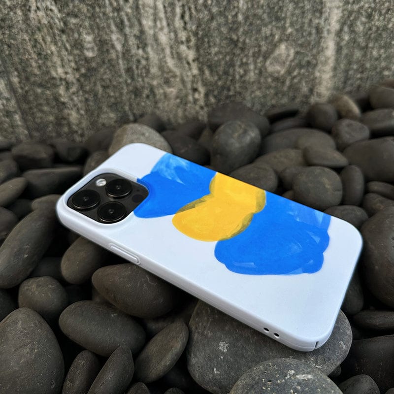 A smartphone encased in an Abstract Expressionist iPhone 16 Pro Max Case - Bold Blue & Yellow Brushstrokes is lying on a bed of smooth gray stones. The phone features triple rear cameras and a metallic frame, with large, textured stones in the background.