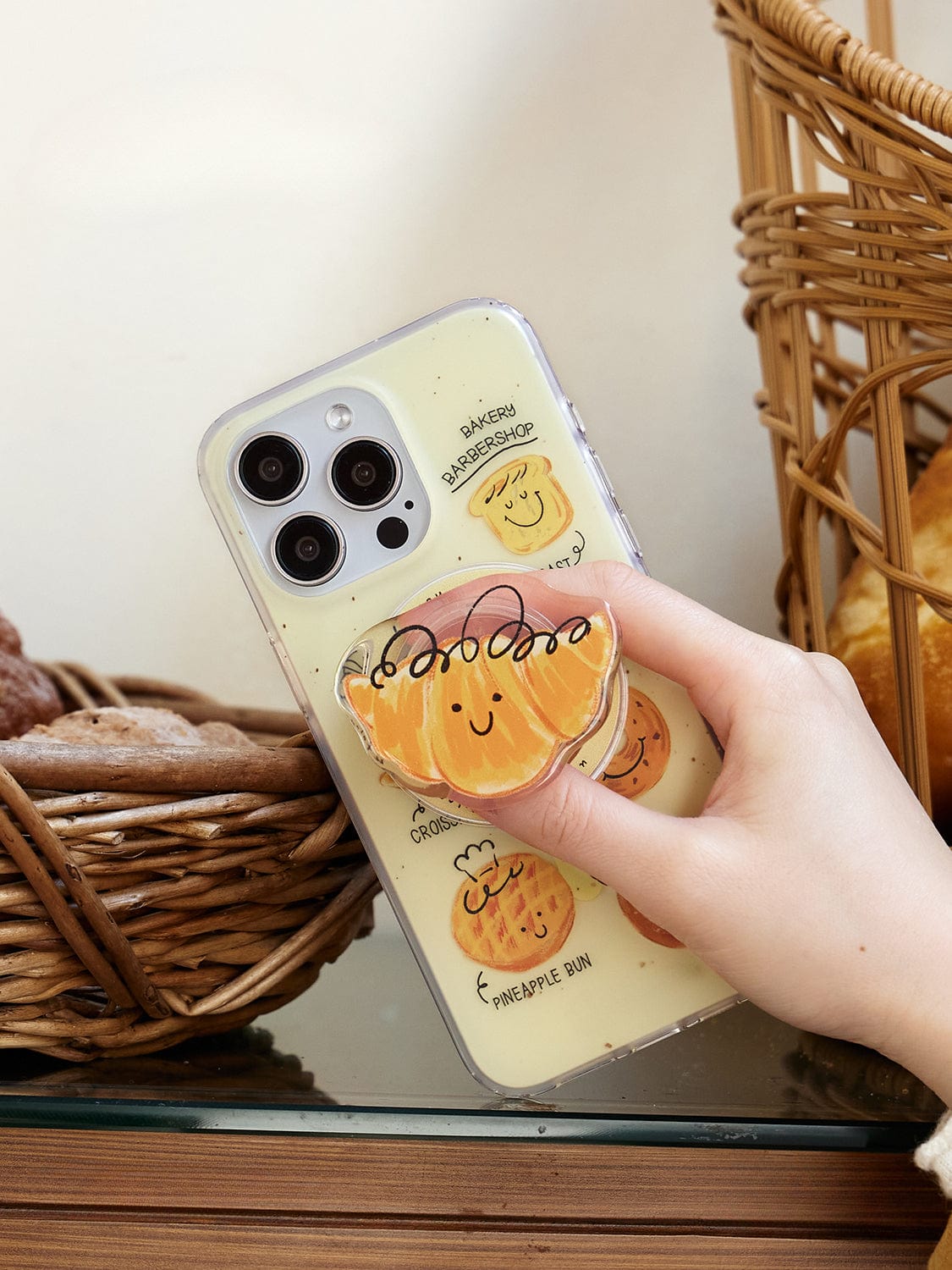 A hand holding the Bakery Friends MagSafe iPhone 16 Pro Max Case, showcasing its transparent design adorned with adorable, smiling pastry illustrations. The phone case features various baked goods designs, including bread rolls and croissants. The background depicts wicker baskets filled with bread.