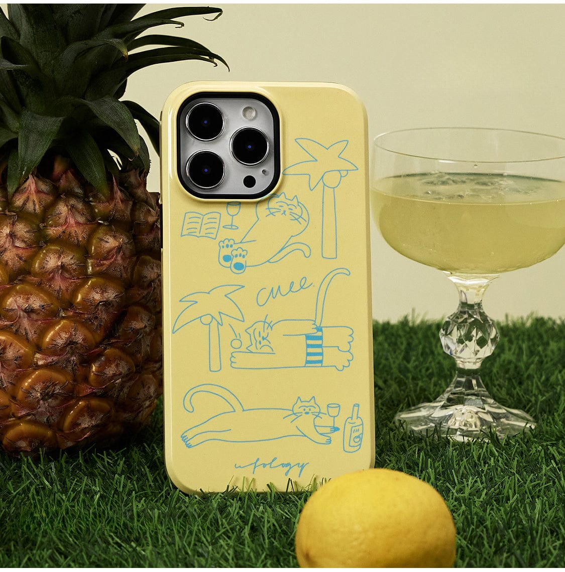 A Tropical Cat MagSafe iPhone 16 Pro Max Case in sunny yellow, featuring playful cat illustrations and a relaxed cat stand, is propped against a whole pineapple. Beside the phone case is a cocktail glass filled with a drink and a lemon placed in the foreground. The scene is set on a grassy surface.