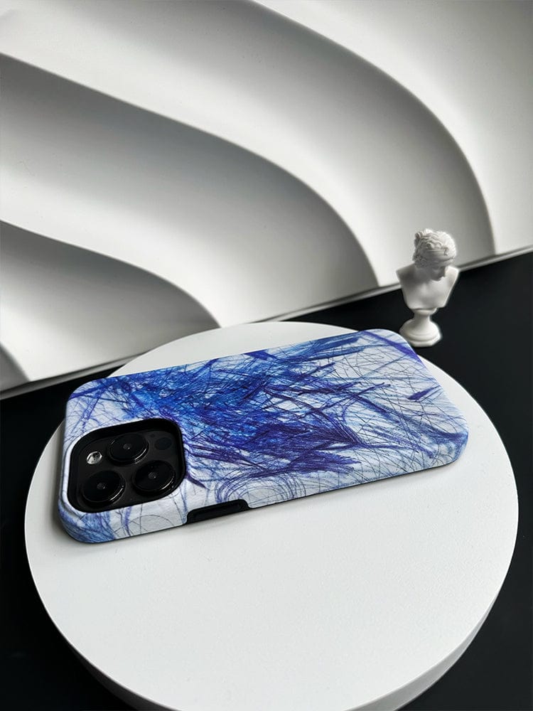 An iPhone 16 Pro Max adorned with the Abstract Blue Scribble case, featuring an artistic design and complete protection, is placed on a round white platform. Nearby stands a small white bust sculpture. White sculpted waves in the background contribute to a modern aesthetic.