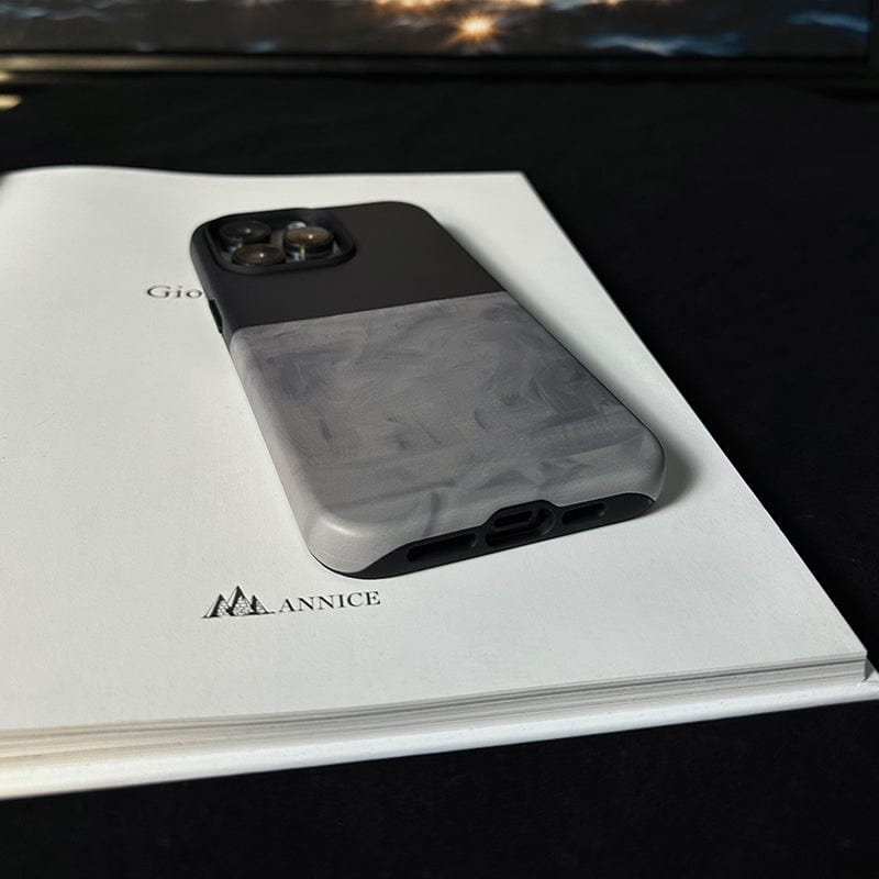 A smartphone with a sleek, Minimalist Two-Tone iPhone 16 Pro Max case in elegant dark green and gray lies face-down on an open book. The book has the text "Gio" and "ANNICE" visible, with a small triangular logo above the word "ANNICE." The background is dark, creating a contrast with the book and phone.