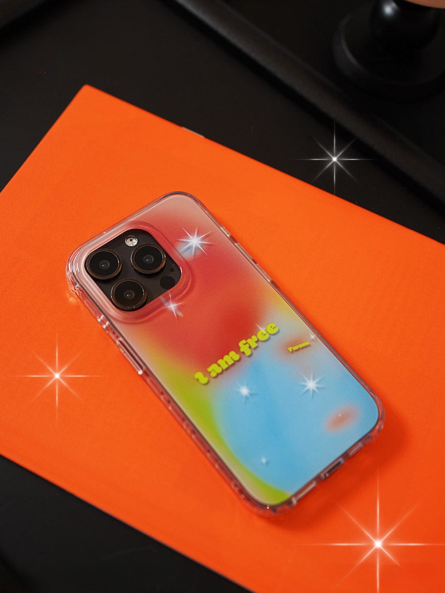 A smartphone with an "I Am Free Gradient iPhone 16 Pro Max Case" lies on an orange surface, showcasing its vibrant colorful gradient design in pink, yellow, blue, and green. The case features the phrase "I am free" printed in yellow letters. Light reflections create star-like sparkles on the reflective all-inclusive protective cover.