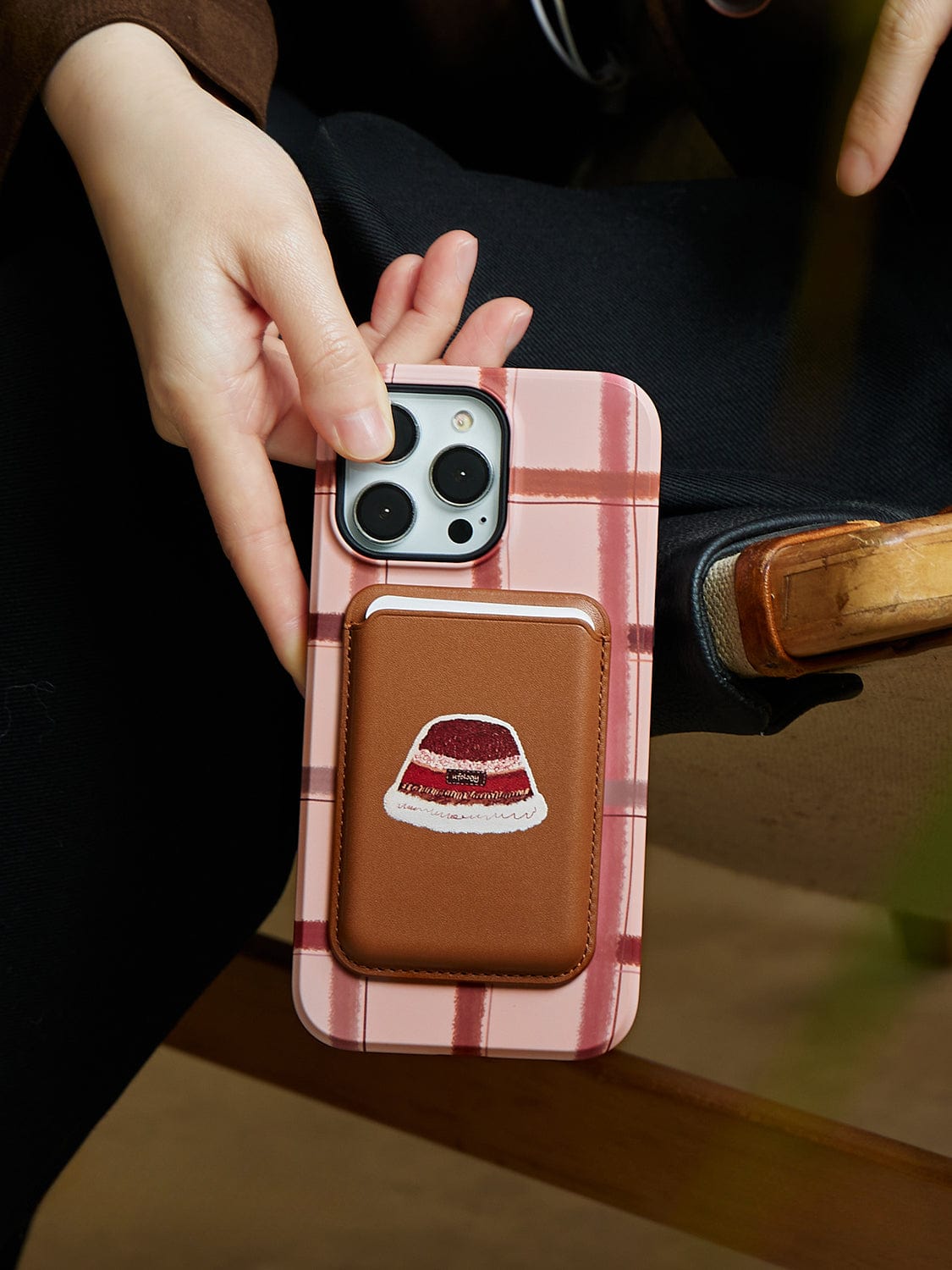 A person holding an iPhone 16 Pro Max with a Cozy Plaid MagSafe case featuring a warm winter hat design and complemented by a brown cardholder attachment.