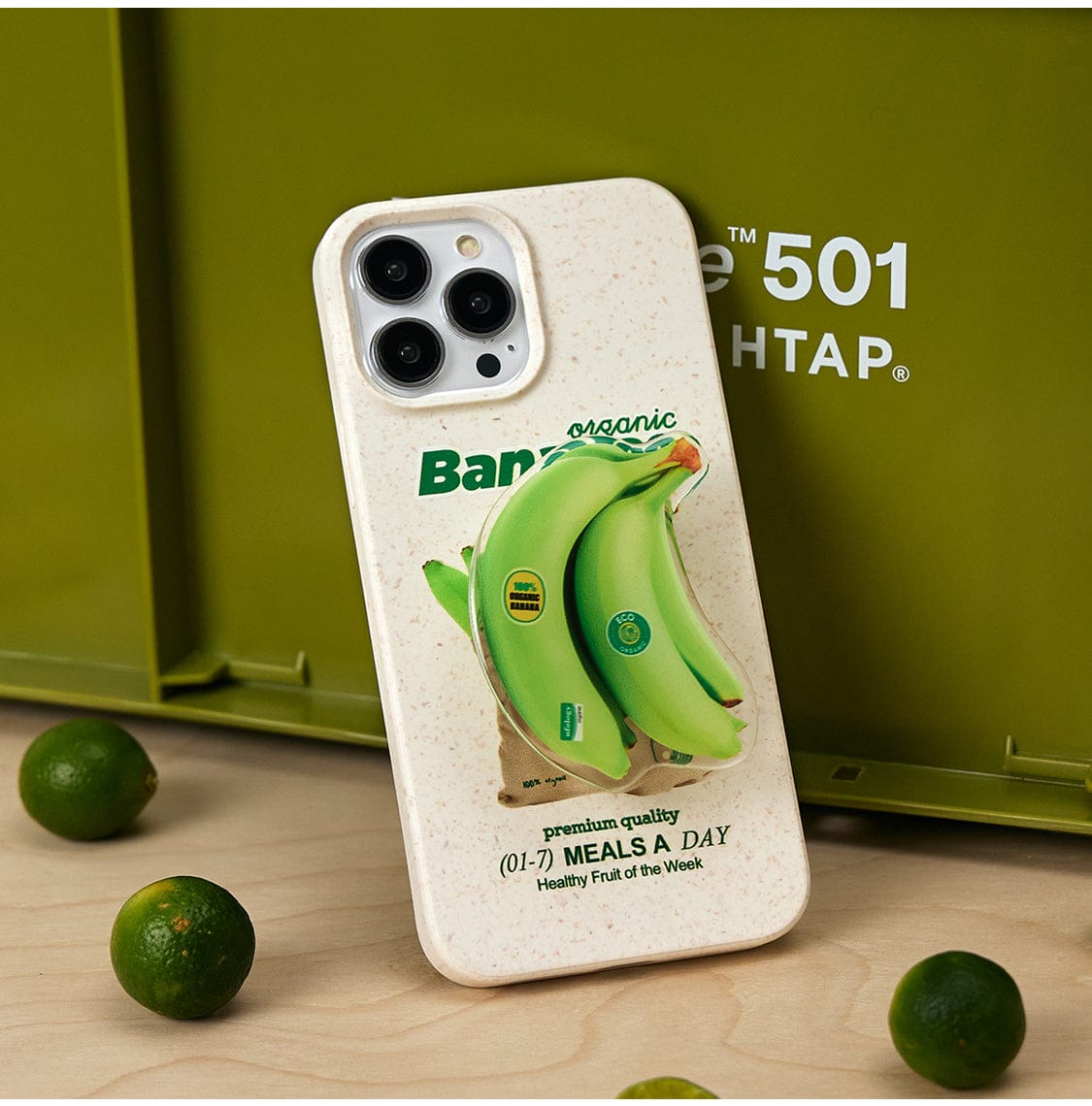 Displayed in the image is a smartphone with an Organic Bananas MagSafe iPhone 16 Pro Max Case. The beige case features an intricate 3D design of two green bananas accompanied by text that reads, "Banana organic," "premium quality (01-7) MEALS A DAY Healthy Fruit of the Week." The background showcases green limes and a green crate, complementing the fun fruit design.