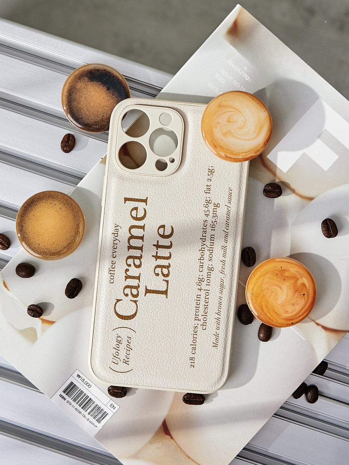 A Caramel Latte MagSafe iPhone 16 Pro Max Case, showcasing a coffee-inspired design with mock coffee stains and drink descriptions, rests on a magazine. Surrounding it are four espresso cups filled with latte and scattered coffee beans on a striped surface.